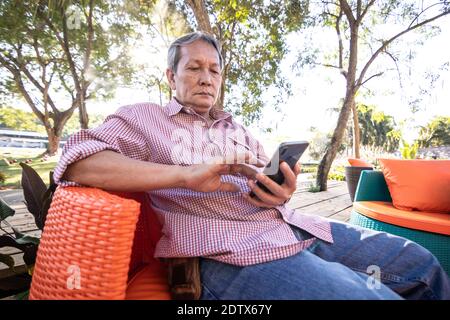 Le persone asiatiche anziane con i capelli grigi usano i telefoni cellulari. Un uomo anziano felice seduto su un comodo divano in giardino. Foto Stock