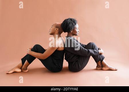 Coppia le ragazze africane di nuovo a indietro che siedono sul pavimento su sfondo marrone in studio Foto Stock