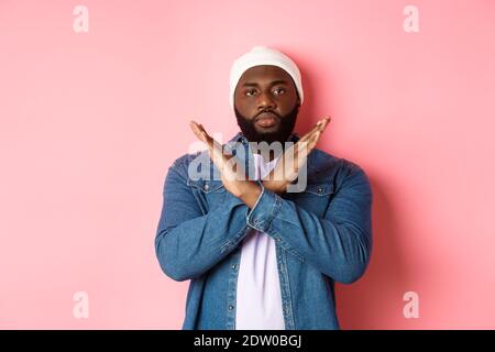 Un uomo afro-americano serio che dice di no, mostrando croce per fermare qualcosa di cattivo, proibisci l'azione, in piedi su sfondo rosa Foto Stock