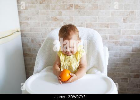 il bambino è seduto su una sedia alta, tenendo un arancione e sorridente Foto Stock