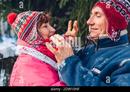 Coppia anziana abbracciata nella foresta invernale. Uomo e donna che camminano all'aperto. Attività invernali. L'uomo stringe il cappello di sua moglie Foto Stock