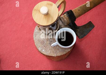 Una pentola di caffè e una pressa francese sul legno tagliato con sfondo rosso tappeto. Un'ascia rimane vicino alla caffettiera e alla pressa francese. Foto Stock