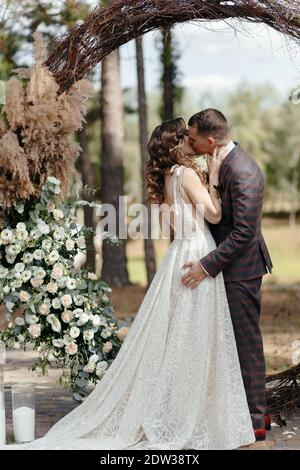 Bride baciante e sposo in piedi davanti all'arco alla cerimonia nuziale. Sposi felici alla cerimonia nuziale Foto Stock