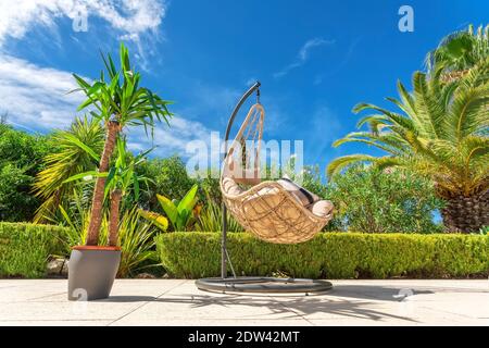 Sedia girevole sospesa per rilassarsi in un giardino di lusso, vista dal basso con un vaso di fiori. Foto Stock