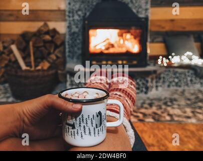 Una donna che tiene una tazza di cacao o cioccolata calda con marshmallows in polvere da un caldo camino in inverno Foto Stock