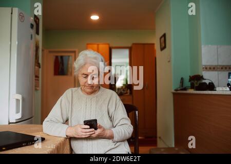 Una donna anziana che scrive messaggi al telefono alla sua famiglia mentre si siede a un tavolo in casa. Comunicazione durante l'isolamento della corona COVID-19 Foto Stock