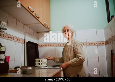 Pranzo di cucina della nonna caucasica in cucina. Una donna ritirata dai capelli grigi accanto ad una stufa da cucina Foto Stock