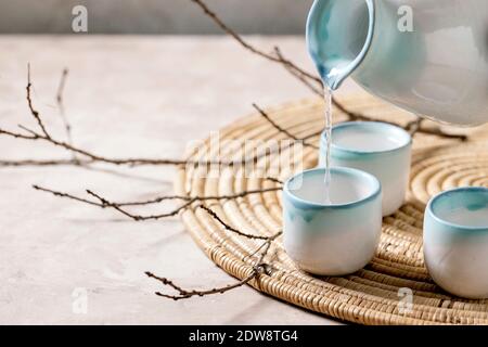 Set in ceramica Sake per bevanda tradizionale giapponese di vino di riso sake versato dalla caraffa in tre tazze, in piedi su tovagliolo di paglia con rami asciutti Foto Stock