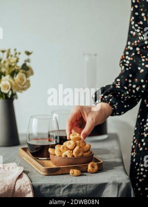 Taralli o tarallini tradizionali con spuntino italiano e vino rosso sul tavolo all'interno. Donna irriconoscibile in abito festivo prende taralli dalla ciotola. Foto Stock