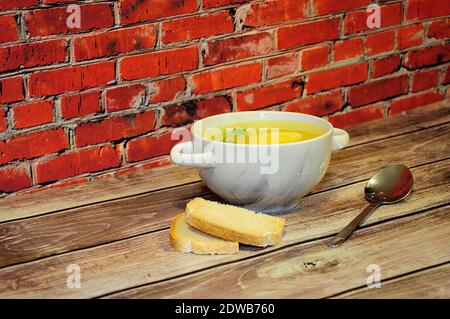 Un piatto profondo di brodo di pollo con uovo e prezzemolo è su un tavolo di legno contro un muro di mattoni, accanto ad un cucchiaio e due fette di pane bianco. Primo piano. Foto Stock