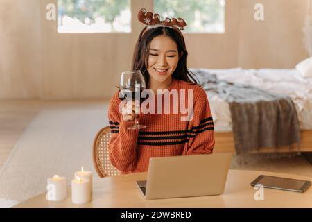Giovane donna asiatica adulta indossa un maglione e orecchie di renna facendo un brindisi in camera interna con un bicchiere di vino e candele sul tavolo. Bella ragazza che usa il laptop videochiamata online durante il festival di Natale Foto Stock
