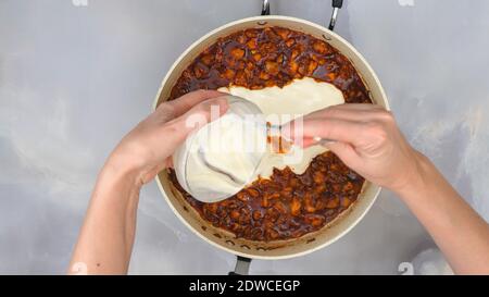 La donna aggiunge panna acida nella padella con carne di pollo marinata tagliata. Foto Stock