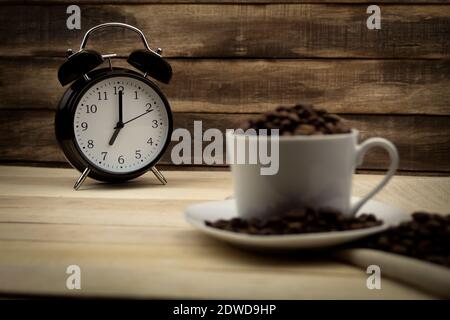 La sveglia mostra le sette. Tazza bianca con grani di caffè sullo sfondo dell'orologio. Legno di fondo Foto Stock