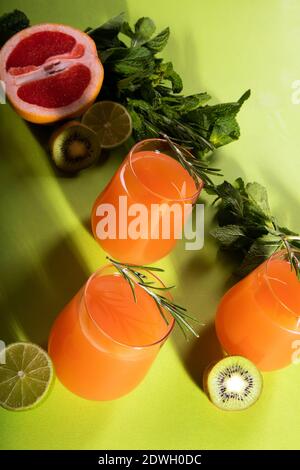 Composizione di bicchieri con succo d'arancia, foglie di arancio, lime, kiwi, rosmarino e menta tagliate su sfondo verde chiaro Foto Stock