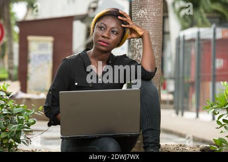 giovane donna che si siede all'aperto e che guarda pensiva al suo lato di spazio di copia. Triste ha sottolineato che lo studente è seduto con un computer portatile sui suoi piedi Foto Stock