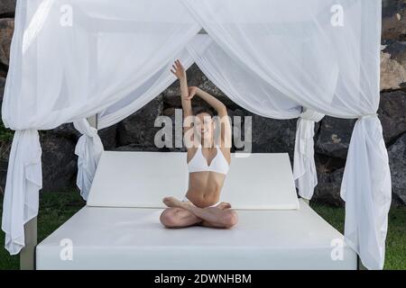 Allegra donna in bikini bianco seduta in Padmasana su baldacchino letto e braccia stretching mentre si pratica yoga e guardare fotocamera Foto Stock