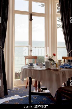 Il sole del mattino illumina un tavolo da pranzo per la colazione con tazze da tè, piatti e posate. In lontananza, l'oceano si estende verso l'orizzonte. Foto Stock