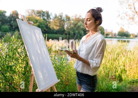 bella ragazza primo piano, artista donna, estate al lago, laghetto fiume, disegni immagine, tavolozza con vernici, pittura bianco tela pittura, un cavalletto Foto Stock