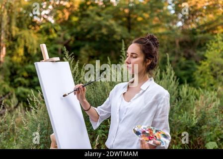 bella ragazza, artista donna, estate nel parco vicino ai cespugli, disegni quadro, tavolozza con vernici, pennello tela bianca pittura, cavalletto stand. Bianco Foto Stock