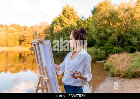 bella ragazza, artista donna, estate al lago, laghetto del fiume, disegna immagine, tavolozza con vernici, vernice bianco tela pittura, un cavalletto stand. Bianco Foto Stock