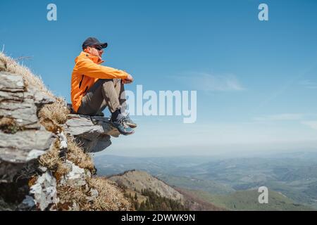 Immagine di concetto di persone attive e turismo europeo. Vestito giacca arancione brillante escursionista in cappellino da baseball e occhiali da sole seduti su scogliera rocciosa godendo di gr Foto Stock
