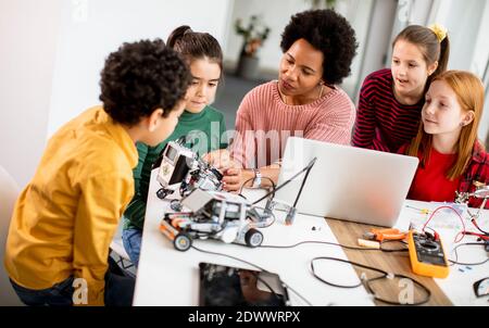 Gruppo di bambini felici con la loro scienza femminile afroamericana insegnante con i giocattoli elettrici di programmazione del laptop e robot alla robotica in aula Foto Stock