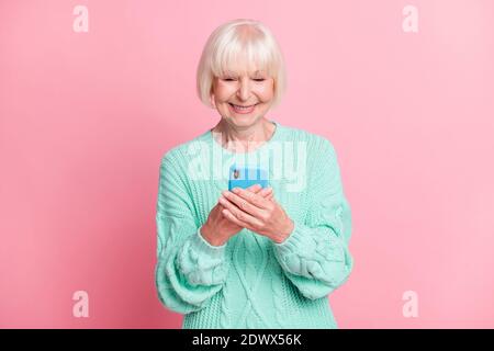 Ritratto fotografico di una vecchia signora allegra che tiene lo smartphone sorridente indossato pullover in teal lavorato a maglia isolato su sfondo di colore rosa pastello Foto Stock
