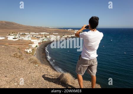 Giovane uomo in piedi sulla scogliera per scattare foto della città costiera di Puerto Calero vicino all'oceano. Turista maschile utilizzando il cellulare per catturare il paesaggio naturale Foto Stock
