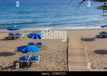 Sentiero in legno che conduce alla riva con amache e ombrelloni blu sulla spiaggia vuota a Puerto del Carmen, Lanzarote. Concetto di ascenza turistica Foto Stock