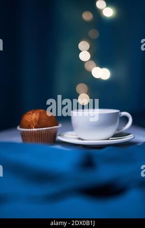 Tazza bianca di caffè con gustoso muffin con luci sfocate su sfondo blu nella caffetteria Foto Stock