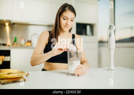 Donna con miscelatore a immersione che fa la polvere di proteine di cioccolato di banana milkshake Smoothie.Adding una paletta di basso siero di latte mix proteico a. agitare dopo un ho Foto Stock