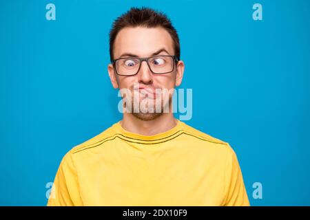Ritratto di giovane uomo con camicia gialla e nerd alla moda vetri isolati su sfondo blu con volto confuso e divertente espressione Foto Stock
