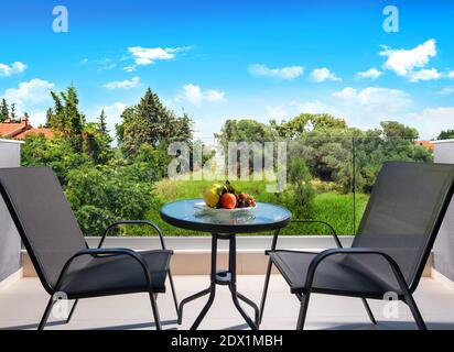 Vista frontale del balcone aperto con vista sul giardino estivo nella foresta di campagna. Terrazza patio con tavolo di vetro, sedie e frutta piatto Foto Stock