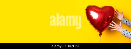 La mano della donna raggiunge il pallone d'aria a forma di cuore su uno sfondo giallo. Banner. Foto Stock
