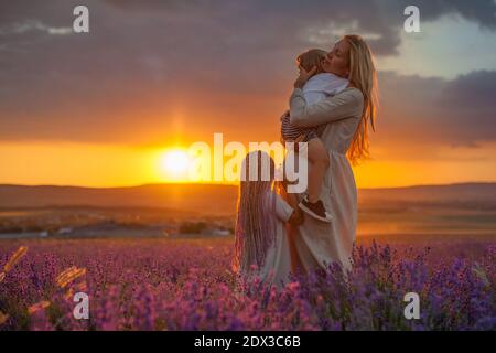 Una giovane madre incinta con molti bambini sta abbracciando suo figlio, sua figlia sta in piedi e guardando il sole che tramonta in un campo di lavanda Foto Stock