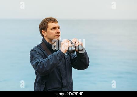 Un giovane uomo su uno sfondo di mare guarda nel distanza e tiene in mano binocoli Foto Stock