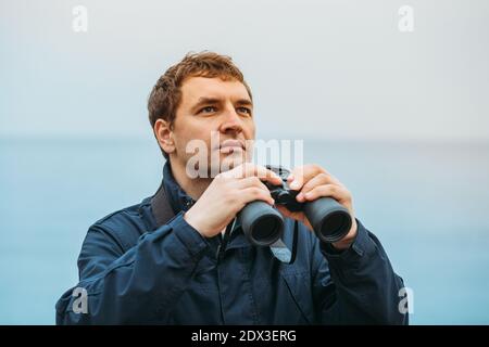 Un giovane guarda qualcosa nel cielo sul mare e tiene in mano binocoli Foto Stock