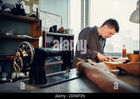 A young shoemaker in his workshop sews leather on a sewing machine, makes original shoes to order. Stock Photo