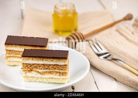 Torta al miele con guarnizione al cioccolato e crema al burro bianco. Dolce di nozze fatto in casa. Servito su un tavolo di legno bianco. Miele in vetro come decorazione. Foto Stock