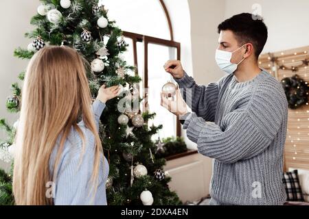 Giovane coppia che decorano l'albero di Natale a casa indossando maschere facciali Foto Stock