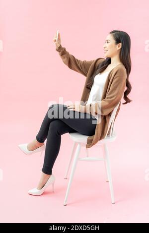 Ritratto a tutta lunghezza di una donna felice in abbigliamento da lavoro presa del selfie sul telefono delle cellule mentre sedendo sulla sedia Foto Stock