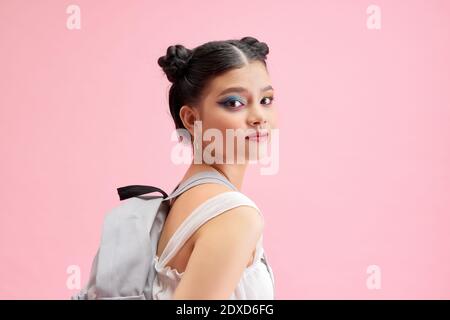 Ragazza scolastica alla moda che tiene uno zaino dietro le spalle e. guardando direttamente la telecamera isolata su uno sfondo rosa brillante Foto Stock