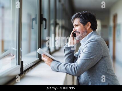 Sorridente professore di sesso maschile che parla al telefono mentre si guarda attraverso la finestra in corridoio all'università Foto Stock