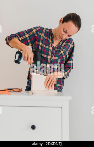 Donna mid adulta che utilizza un trapano elettrico mentre lavora in un armadietto a casa Foto Stock