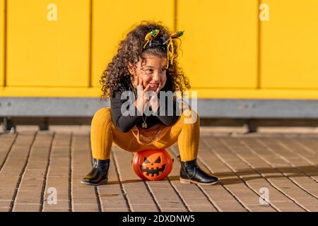 Ragazza che indossa la fascia seduta sul vaso di fiori di Halloween contro il giallo parete Foto Stock
