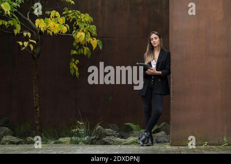 Giovane bella imprenditrice femminile che tiene un tablet digitale mentre si appoggia parete marrone Foto Stock