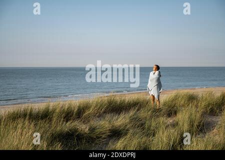 Giovane donna avvolta in scialle in piedi sulla spiaggia Foto Stock