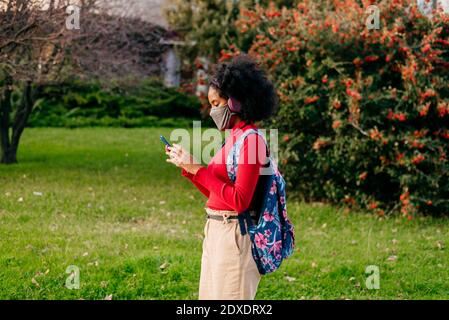 Studente che indossa le cuffie e la maschera protettiva per il viso utilizzando lo smartphone in piedi all'aperto Foto Stock