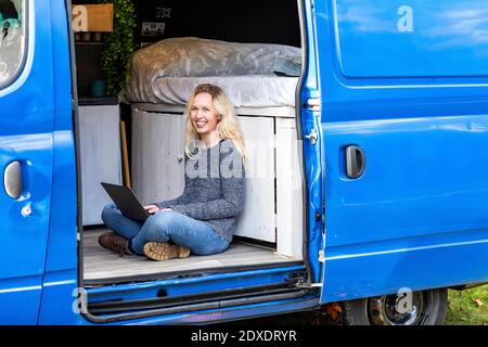 Donna felice con il laptop seduto alla porta del camper van Foto Stock