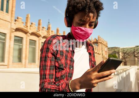 Afro giovane donna che indossa una maschera sanitaria rosa mentre si usa Smart Telefono durante COVID-19 Foto Stock